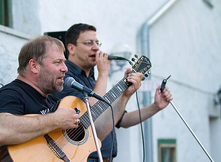 Zwei Männer singen bei einem Konzert in ein Mikrofon. Einer der beiden spielt Gitarre.