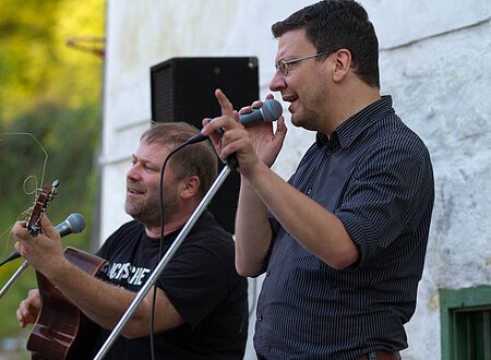 Zwei Männer singen bei einem Konzert in Mikrofone. Einer der beiden spielt Gitarre.