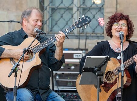 Ein dunkel gekleideter Herr mit Gitarre in der Hand und eine dunkel gekleidete Frau mit Brille, ebenfalls mit Gitarre. Vor beiden steht ein Notenständer und zwei Mikrofone. Im Hintergrund sieht man einen Verstärker stehen und ein vergittertes Fenster.