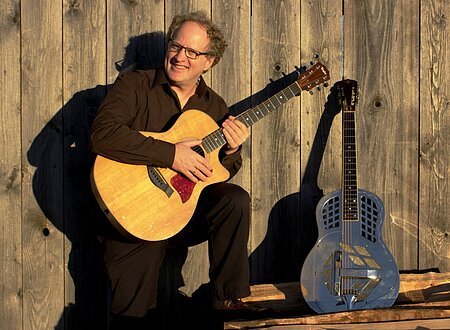 Ein dunkel gekleideter Mann mit Brille und Gitarre in der Hand. Eine weitere Gitarre lehnt an einer Holzwand.