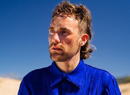 Ein Mann mit längerem Haar und blauem Hemd, im Hintergrund blauer Himmel.