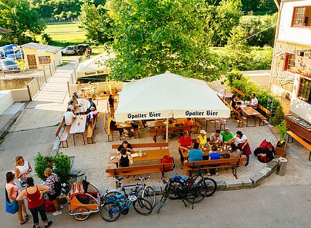 Biergarten in der Aktivmühle Solnhofen Biergarten mit mehreren Tischen, Sonnenschirmen „Spalter Bier“, Menschen sitzen und stehen, Fahrräder davor.
