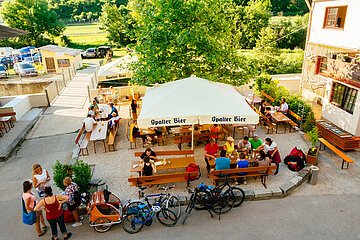 Biergarten mit mehreren Tischen, Sonnenschirmen „Spalter Bier“, Menschen sitzen und stehen, Fahrräder davor.